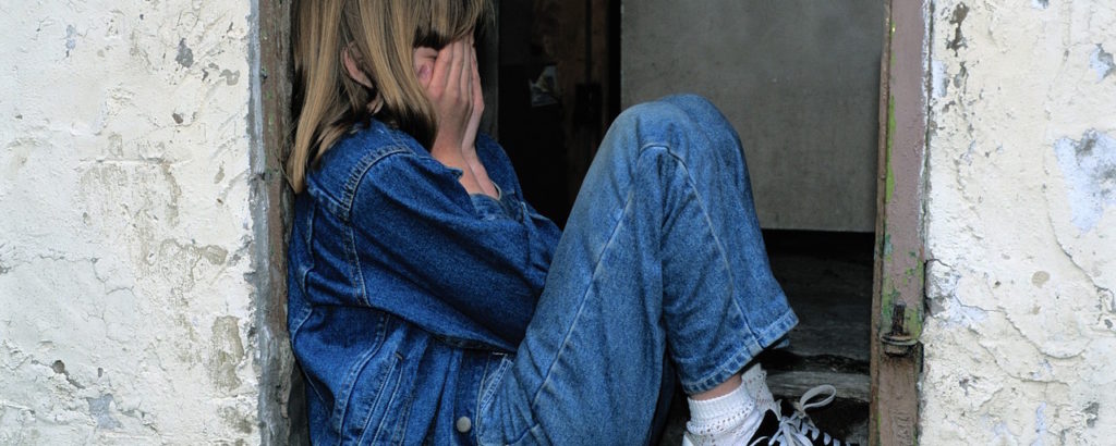 A child sitting in a doorway with their hands covering their face, presumably crying.