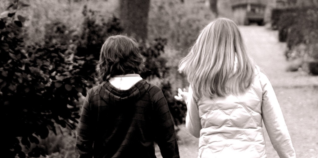 A black and white photo of two kids walking along a path. The photo is taken from behind, so only the backs of them are shown.