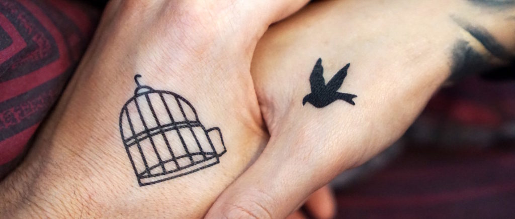 Two people holding hands. One hand has a tattoo of a bird on it, and the other hand has a tattoo of an empty birdcage.