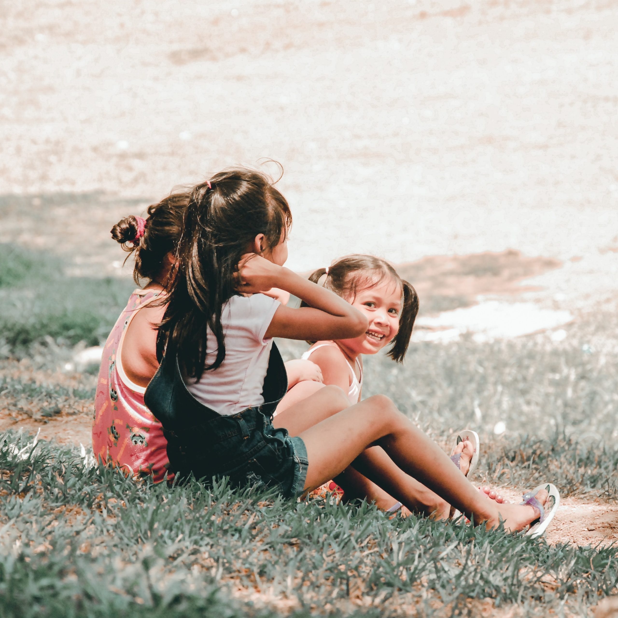 Three kids sit on the grass. Two of them are facing away, but one is looking directly at the camera smiling.