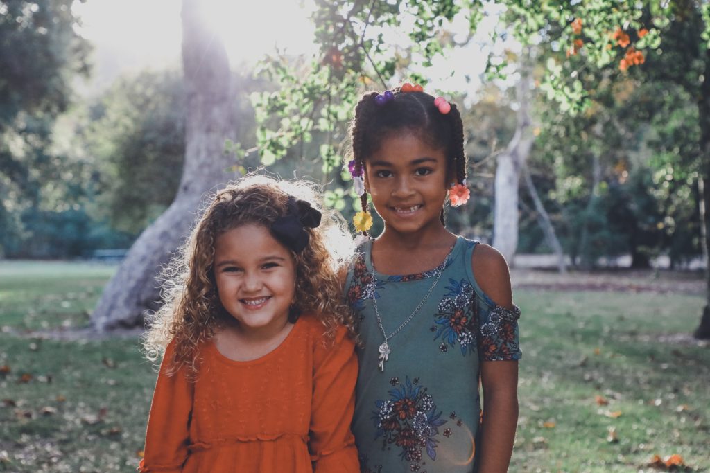 Two young girls standing next to each other, smiling at the camera. They are standing outside with trees and the sun behind them.
