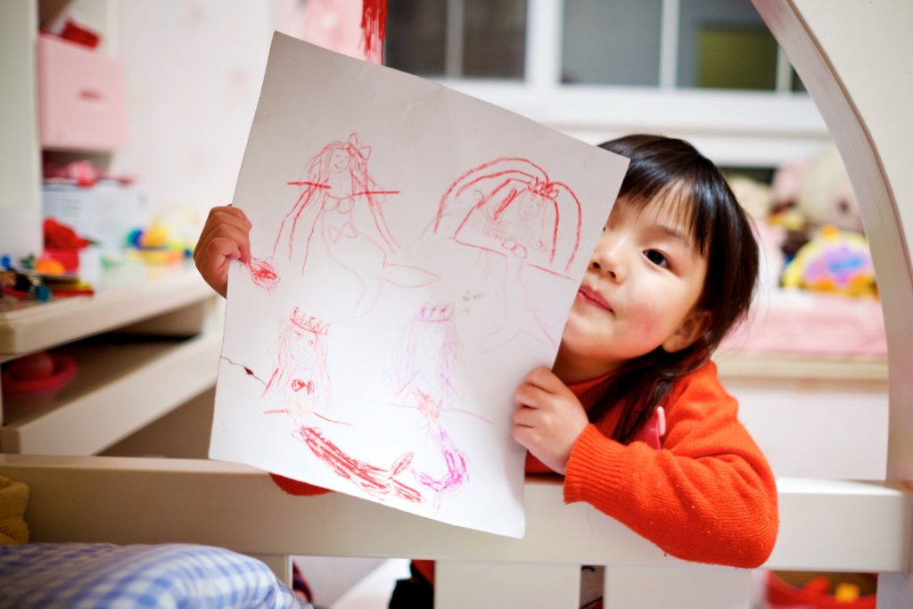 A young girl proudly holding up a picture they drew with crayons.